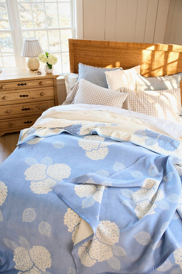 Blue blanket with cream hydrangea flower and leaf pattern draped over a bed with pillows.2
