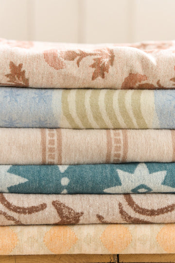 Stack of six folded lightweight blankets in tan, cream, blue, and teal with stripe and celestial patterns.6