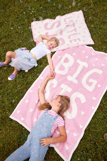 Pink mini blanket with "Little Sis Big Sis" text and white hearts, displayed on grass with two children.6