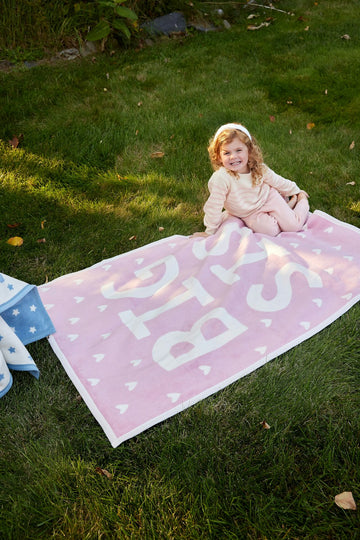 Pink midi blanket with white text and heart pattern, displayed on grass with child sitting beside it.7