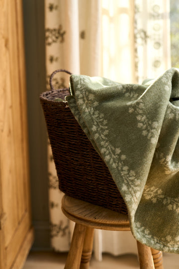 Green and white patterned lightweight blanket draped over a woven basket on a wooden chair near a window.7