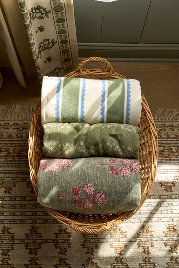 Three folded blankets stacked in wicker basket: top striped in cream, blue and green; middle solid green; bottom sage green w7