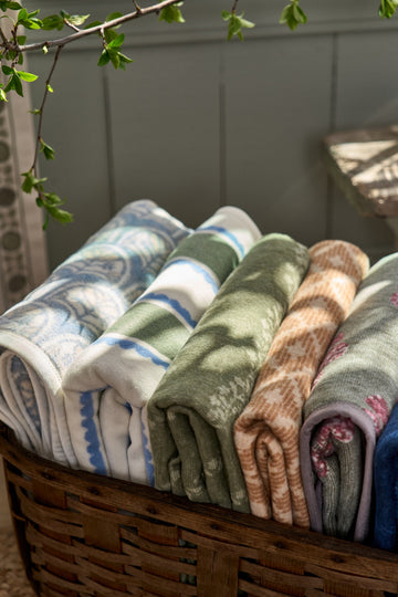 Rolled lightweight blankets in blue, white, green, tan, and gray tones stacked in a woven wooden basket.5