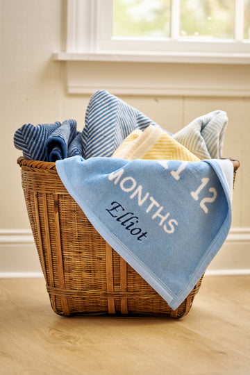 Light blue blanket with "12 MONTHS" and "Elliott" text draped over wicker basket containing striped baby blankets.7