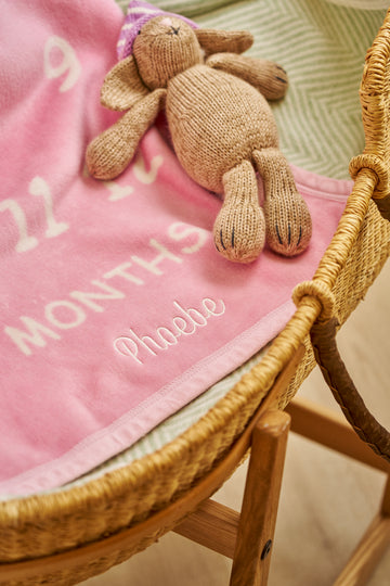 Pink blanket with embroidered text and a tan knit teddy bear with purple hat in a wicker bassinet.5