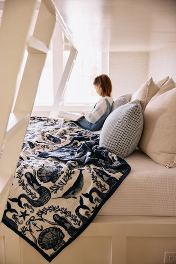 Cream and navy blanket with mermaid and seaweed pattern draped over a couch near a window.7