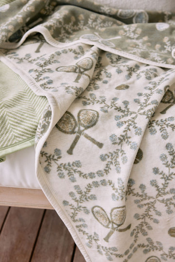 Cream blanket with tennis racket and floral pattern, white binding, layered with green striped fabric.6