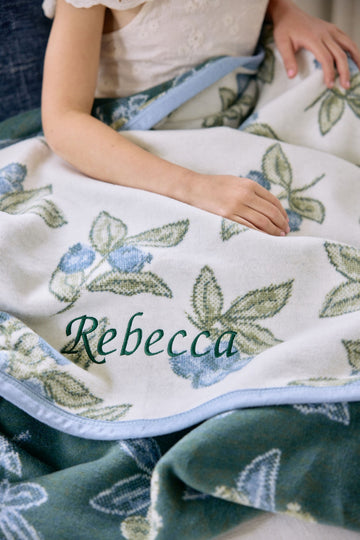 Cream blanket with blue blueberries and green leaves pattern, personalized with "Rebecca" embroidered text.2