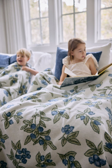 Cream-colored blanket with blue blueberries and green leaves pattern, draped over two children sitting on a bed.6