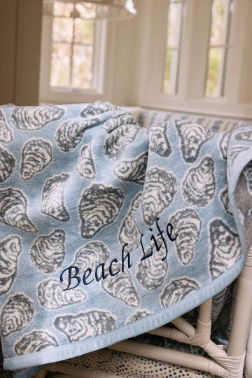 Light blue blanket with gray oyster shell pattern and "Beach Life" text draped over wicker chair.2