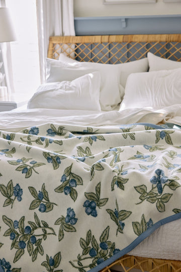 Cream blanket with blue blueberries and green leaves pattern draped over white pillows on a bed.4