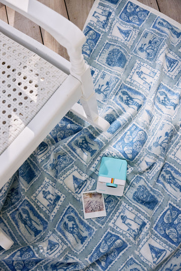 Blue and white blanket with stamp-patterned squares displayed with white chair and instant camera printer.7