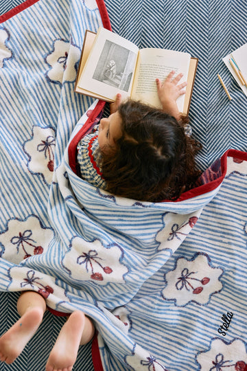 Blue and white striped midi blanket with cherry motifs and red trim, displayed flat with child reading on it.7
