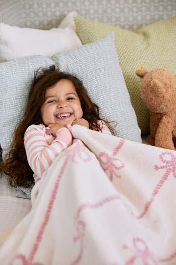 Child wrapped in cream blanket with pink bow pattern, lying on pillows with stuffed animal nearby.3