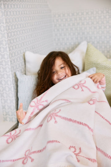 Child wrapped in cream blanket with pink bow pattern, resting against pillows on bed.2
