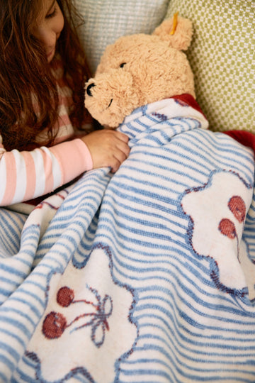 Midi blanket with blue stripes and red cherry design, held by child with tan plush bear on lap.6