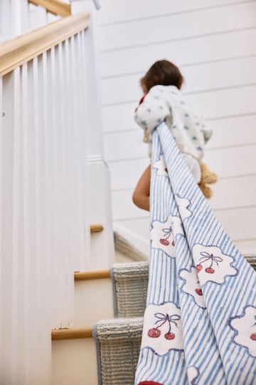 Midi blanket with blue striped and cream patterned sections featuring red cherry designs, draped over child on staircase.3