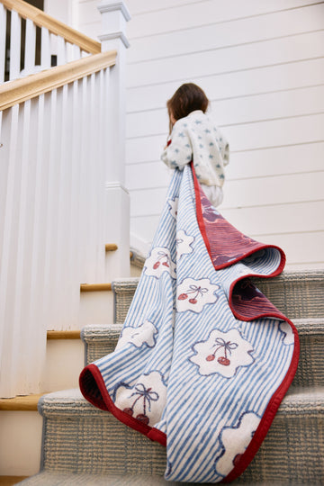 Child wearing blanket with blue stripes, cherry and bow print, red binding, draped over stairs.2