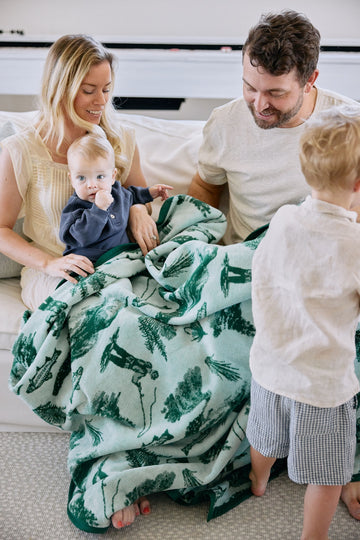 Cream blanket with green fish and botanical print, shown draped over a family with young children on a sofa.4