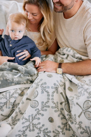 Two adults and a toddler sitting on a cream blanket with gray leaf and circular pattern design.3