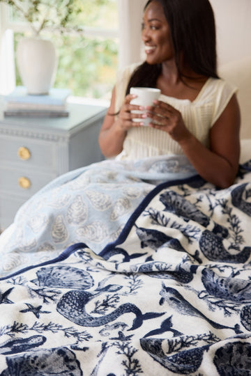 Cream and navy blanket with ocean-themed siren and fish pattern, navy binding, displayed on bed.4
