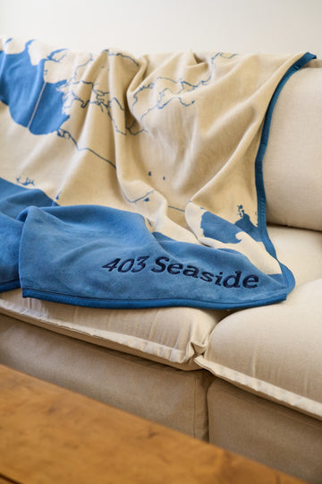 Cream and blue blanket with map design draped over a beige couch, showing "403 Seaside" text on blue binding.6