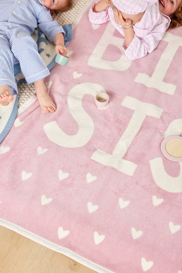 Pink blanket with white "Big Sis" text and heart pattern, shown with two children and cups on it.2