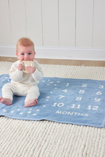 Blue milestone blanket with white numbers, stars, and "months" text, displayed with infant on white textured surface.6