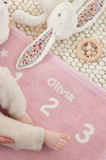 Pink milestone blanket with white embroidered numbers, white plush bunny toy with floral ears, wooden teething ring, and crea5