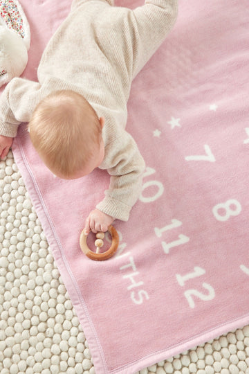 Infant on pink blanket with white milestone text and numbers, holding wooden rattle.4