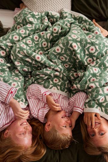 Green and cream knit blanket with floral and botanical print, draped over three children in pink striped clothing.9