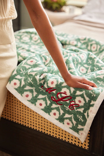 Green and cream blanket with pink floral pattern and red embroidered text on corner, draped over lap.10