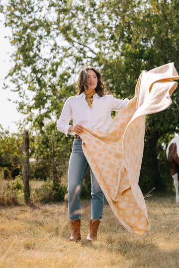 Cream and gold patterned lightweight blanket held up by woman outdoors with trees in background.2