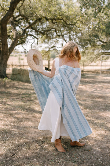 Woman wearing blue and white striped lightweight blanket draped over her shoulders outdoors under a tree.3
