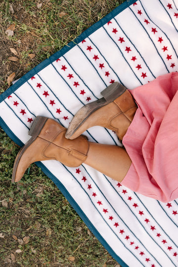 White blanket with red stars and navy stripes, displayed on grass with person wearing brown boots and pink fabric.4