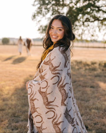 Woman wearing cream and tan patterned lightweight blanket in a pasture with horses and trees in background.4