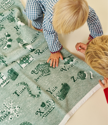 Light green blanket with dark green alphabet letters and Christmas designs, displayed with two children on a cream background2