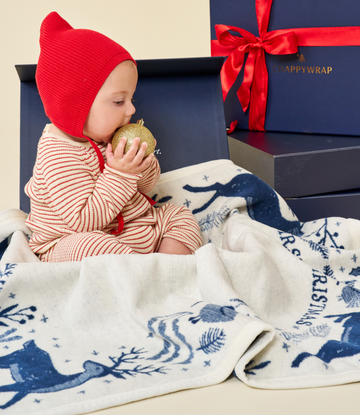 Cream and navy blanket with reindeer and pine tree pattern, displayed on infant wearing red bonnet.3