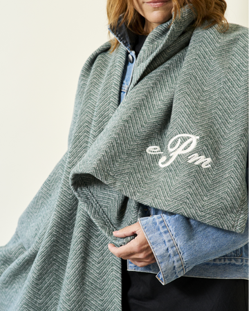 Woman wearing sage green herringbone-patterned shawl with white embroidered logo over blue shirt.8