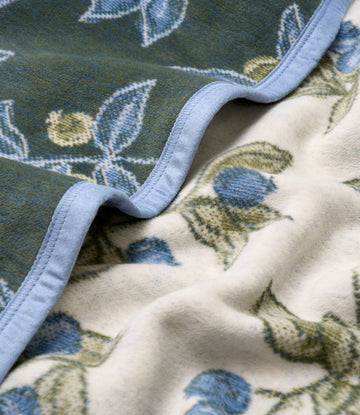 Folded blanket with blue berries and green leaves woven pattern on dark green and cream sides, blue binding edge visible.5