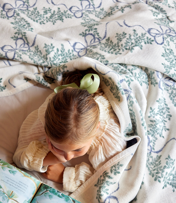 Cream blanket with blue bows and green boxwood leaf pattern, draped over child reading.3