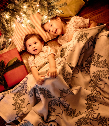 Cream blanket with dark boxwood wreath pattern displayed over two toddlers on wooden floor with holiday decorations.6