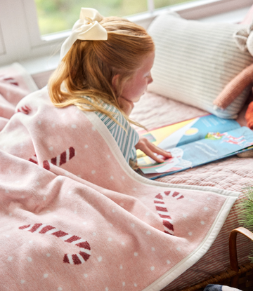 Pink blanket with red candy cane stripes and white dots, child wearing it while reading on a couch.3