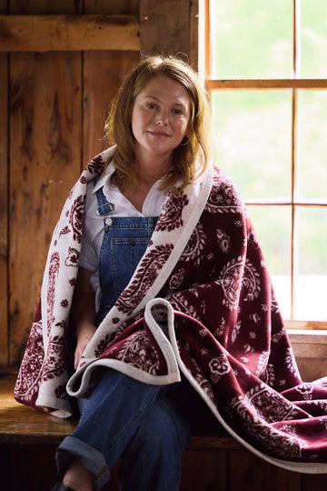 Burgundy and cream paisley blanket with white trim draped over a person's shoulders by a window.2