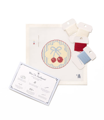 Needlepoint kit with cream canvas showing red cherries with blue bow, instruction booklet, and four spools of embroidery thre3