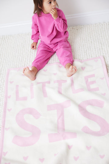 Child in pink outfit sitting on white blanket with pink "Little Sis" text and heart pattern.2