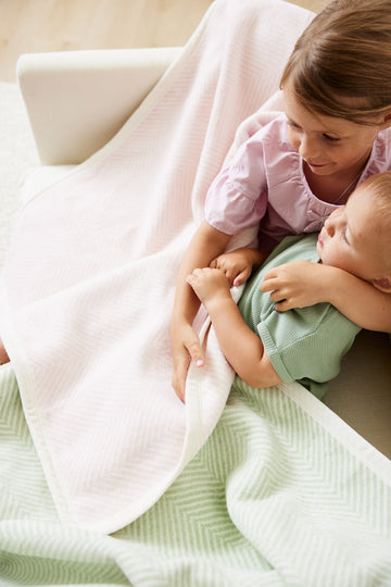 Pink herringbone mini blanket draped over child holding infant in white crib.2
