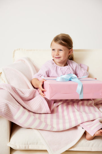Child sitting on a cream chair wrapped in a pink and white striped mini blanket while holding a pink gift box.6
