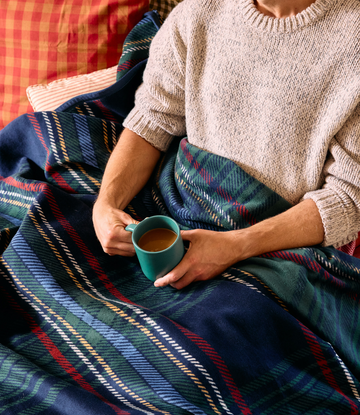 Person wrapped in tartan blanket with navy, green, red and yellow plaid pattern holding a teal mug.2