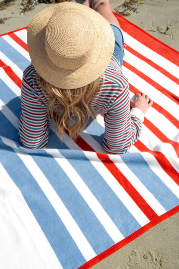 Red, white and blue striped blanket laid on sand with person wearing straw hat sitting on it.2